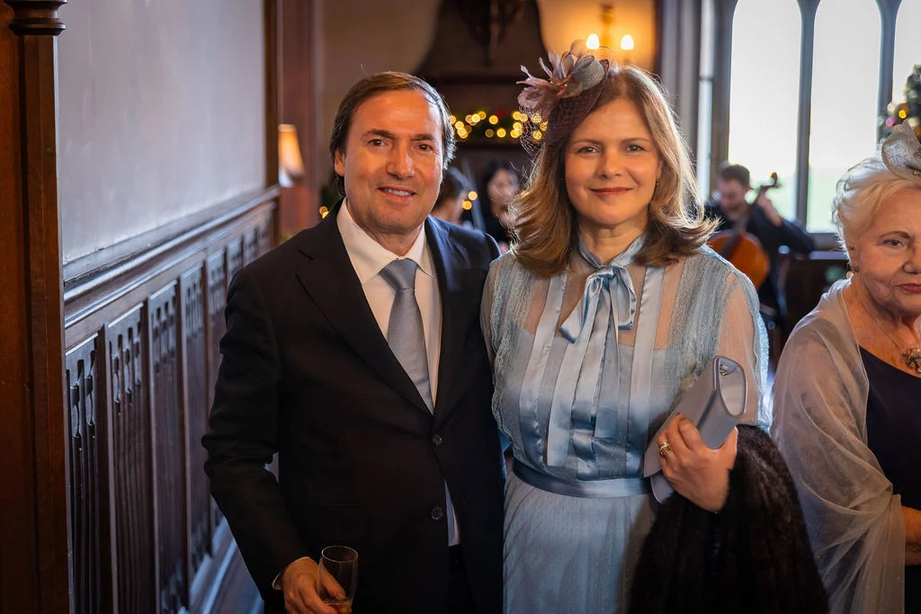 A well-dressed couple poses warmly inside an elegant wood-panelled venue during a wedding celebration.