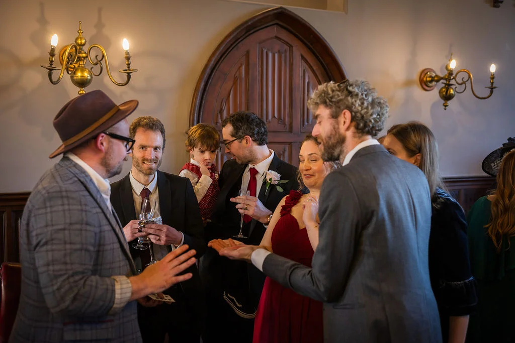A candid shot of wedding guests mingling indoors, watching a magician perform a close-up trick near a wooden archway.