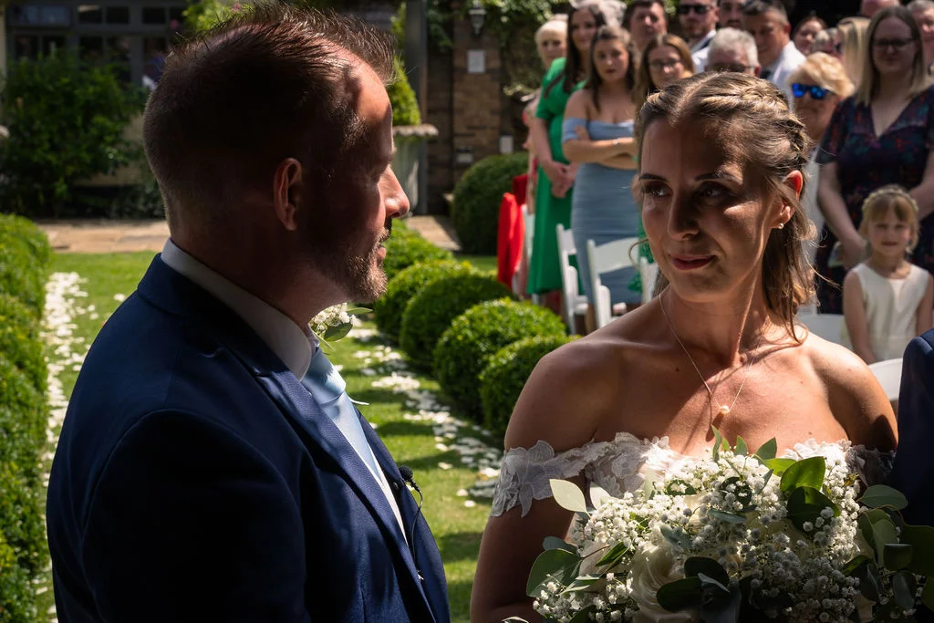 A groom in a navy suit looks adoringly at his bride during their sunlit outdoor wedding ceremony as she holds a lush white bouquet.