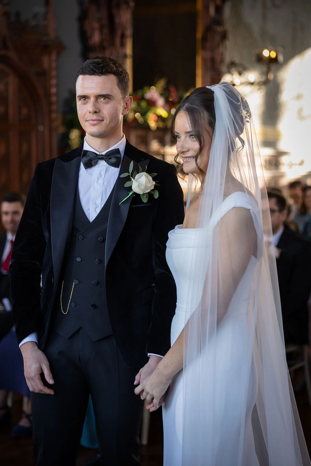 A newlywed couple stands together during their wedding ceremony, with the groom wearing a black velvet tuxedo and the bride in an elegant gown with a veil.