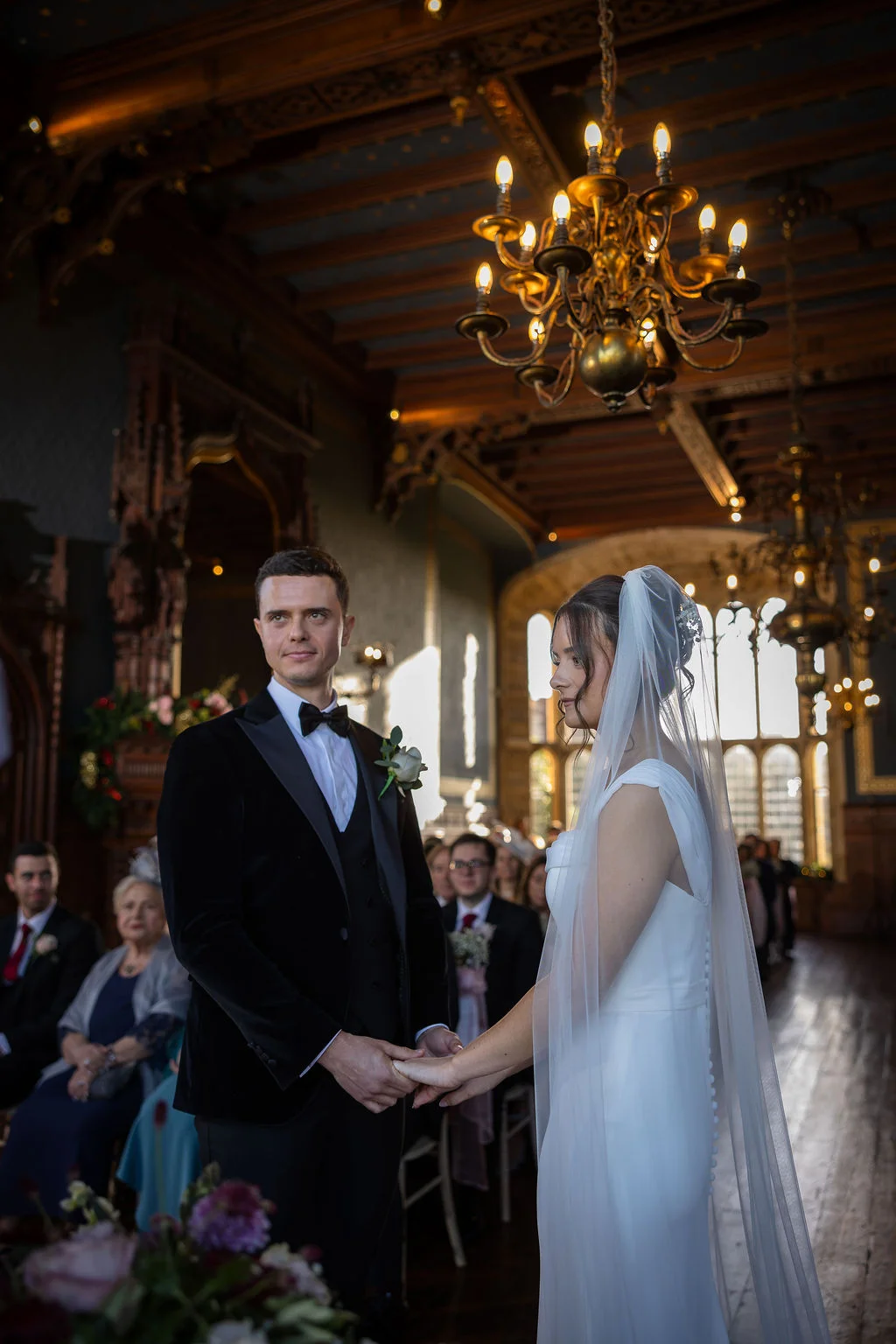 A newlywed couple holds hands during their ceremony inside a historic hall featuring ornate wood panelling and a grand chandelier.