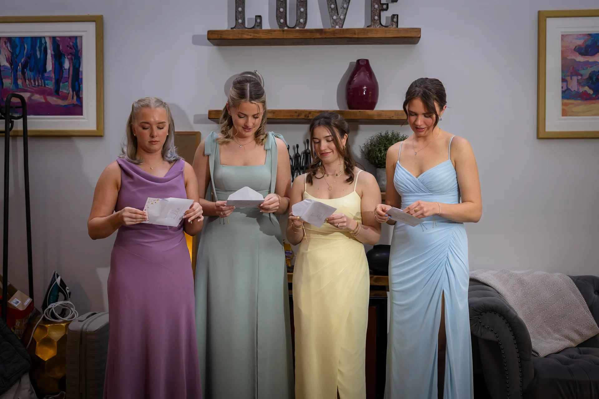 Four bridesmaids in pastel-coloured gowns stand indoors while reading handwritten letters during wedding morning preparations.