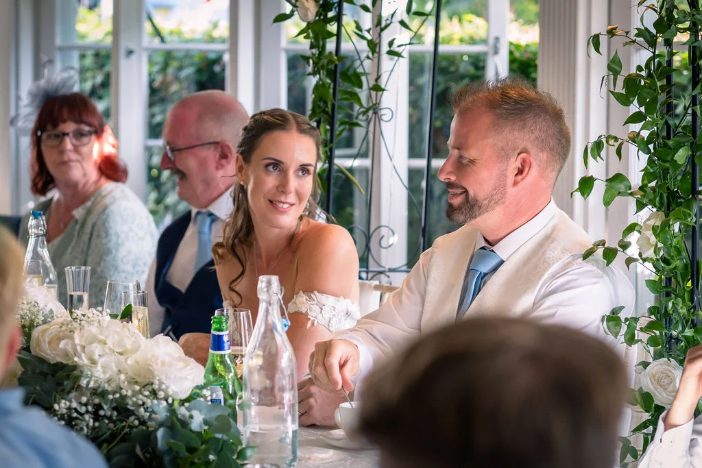 A smiling bride and groom sit at the top table during the wedding breakfast, surrounded by white floral arrangements and guests in a bright, light-filled venue.