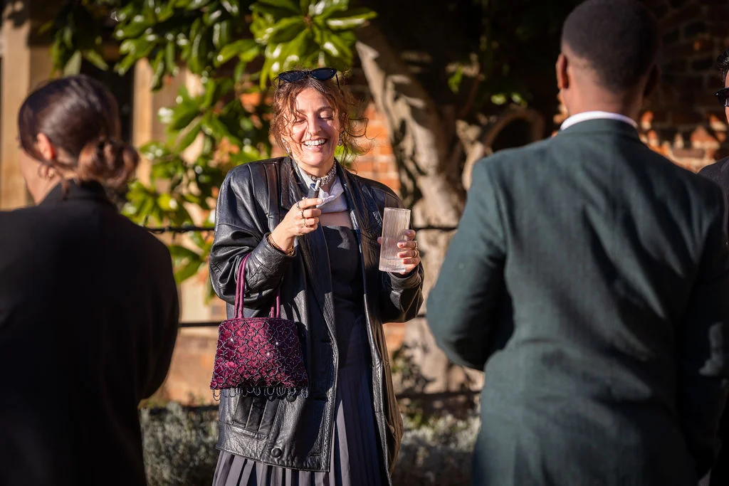 A laughing guest wearing a black leather jacket and holding a glass and beaded bag socialises outdoors during a sunny wedding reception.