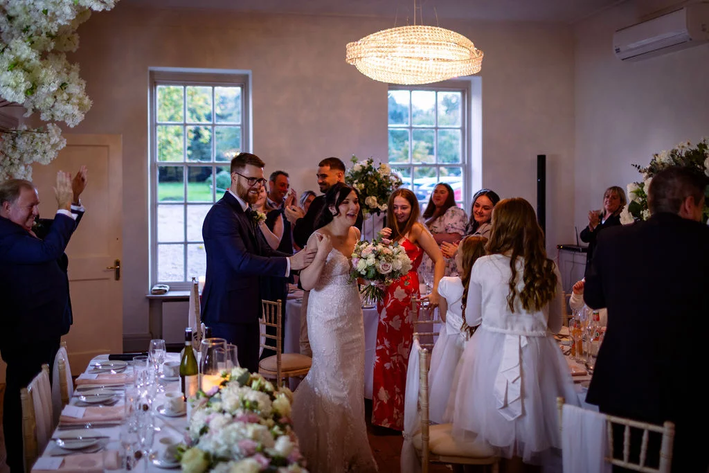 The newly married couple enter the wedding reception venue to applause from seated and standing guests gathered around long banquet tables.