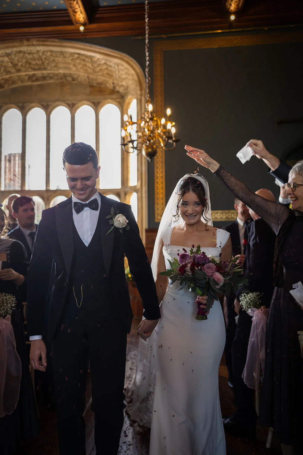 Newlyweds walk down the aisle hand in hand amidst a shower of confetti inside a historic hall with large arched windows and a glowing chandelier.