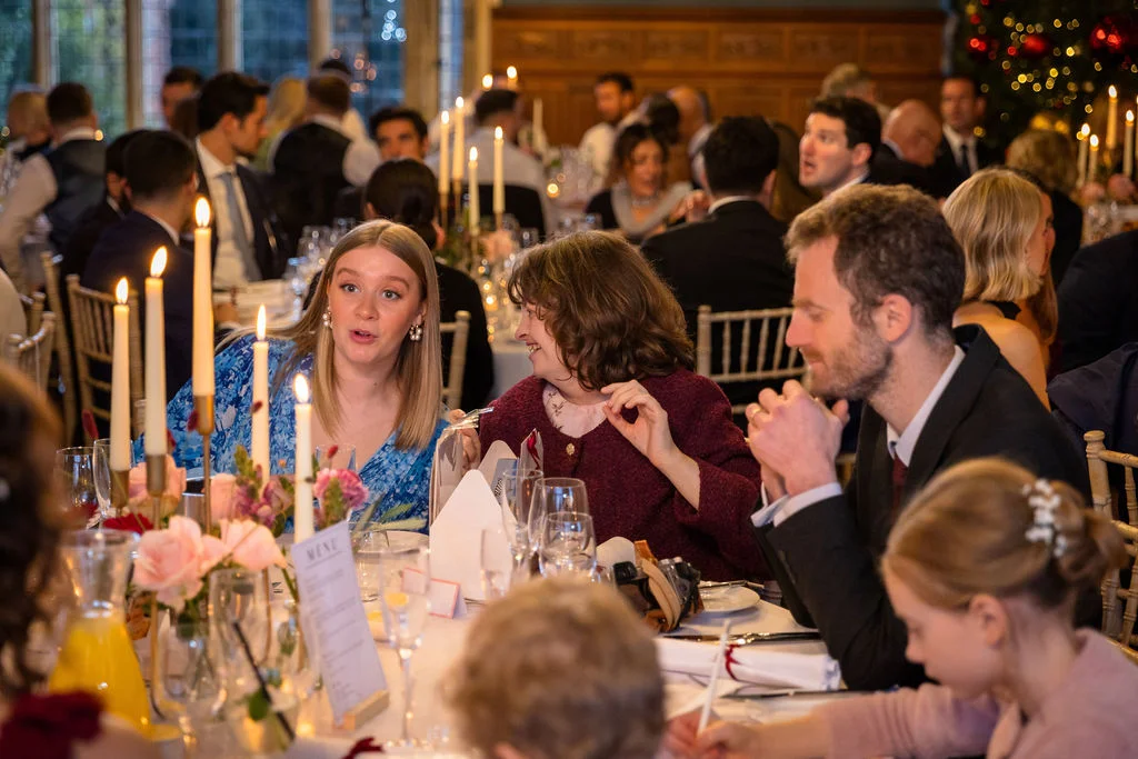 Wedding guests enjoy animated conversation and dinner at a candlelit banquet table during a stately home wedding reception.