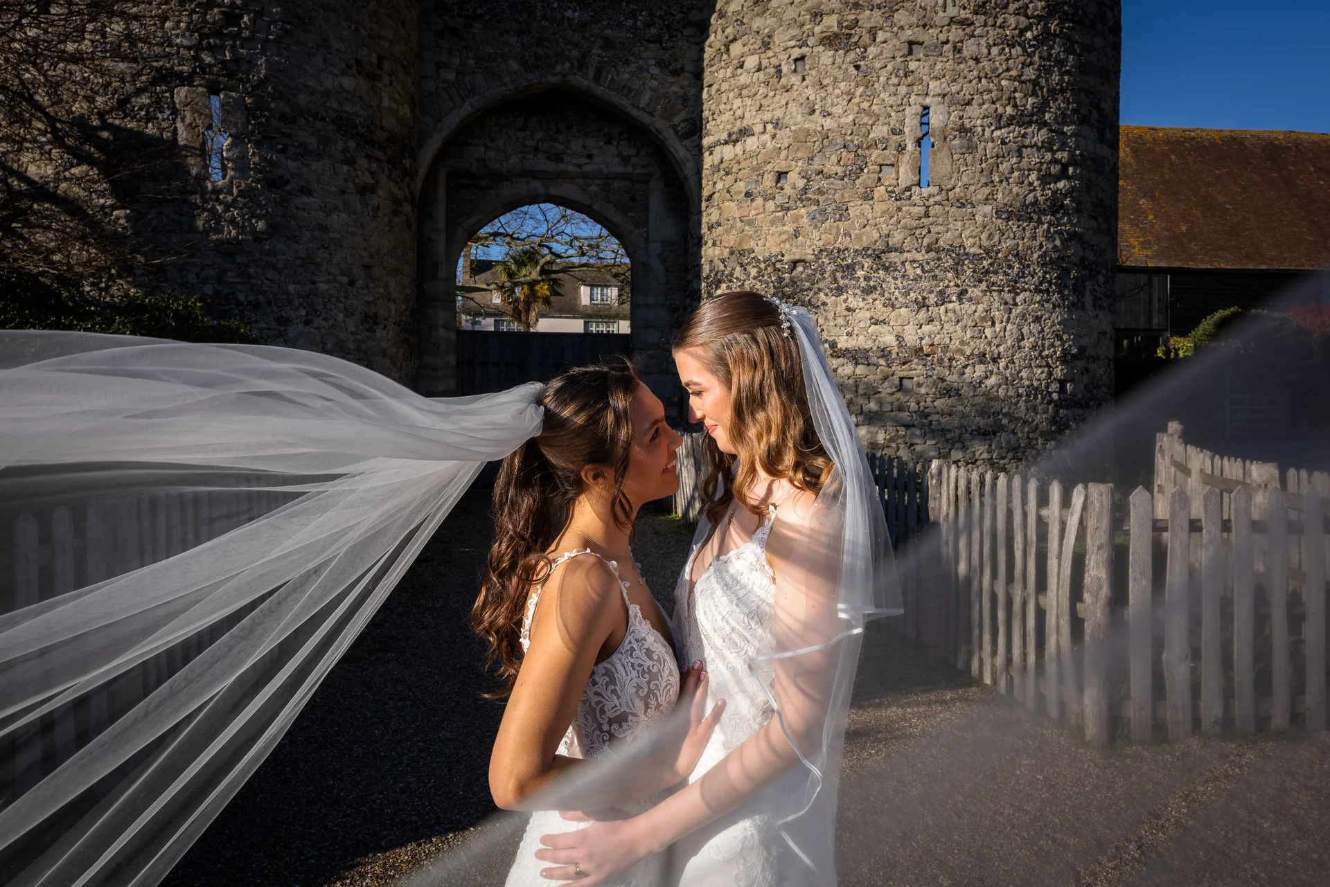Two brides in lace wedding dresses embrace lovingly in front of a historic stone castle archway as their veils billow dramatically in the wind.