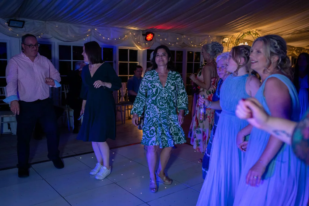 Guests enjoy dancing together on an illuminated dance floor inside a marquee decorated with draped fabric and fairy lights during the evening reception.