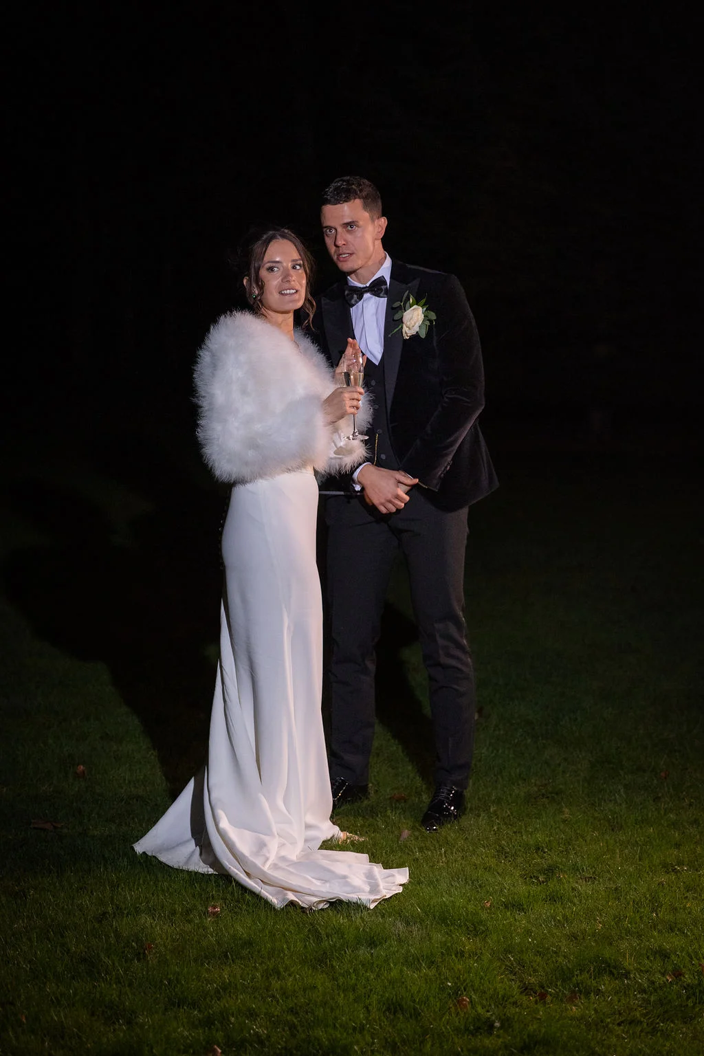 A newlywed couple stands outdoors on grass at night, with the bride wearing a white crepe gown and a feather trim stole while holding a champagne coupe, and the groom in a black velvet tuxedo with a bow tie.