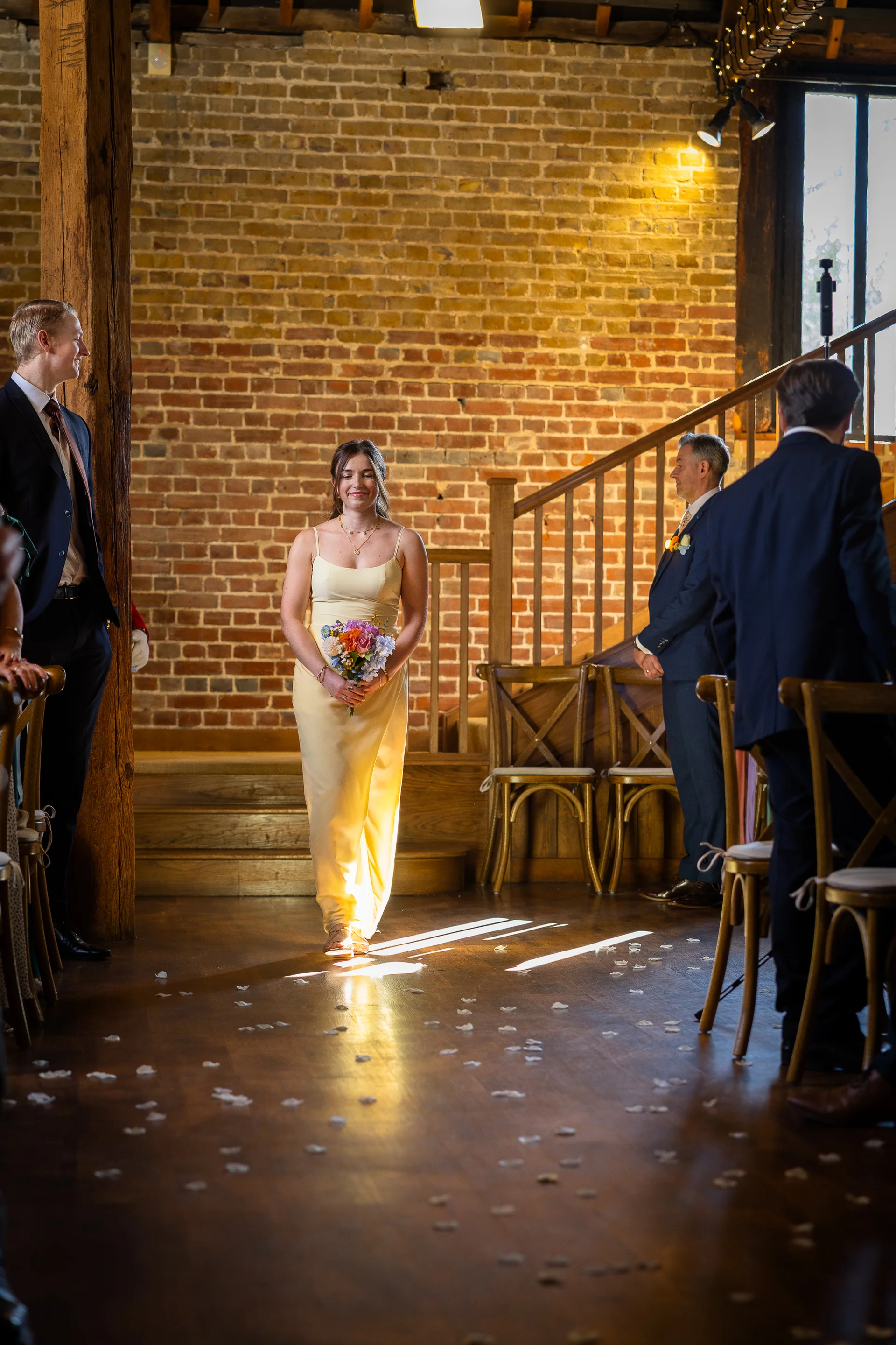 A smiling bride in a yellow dress holding a colourful bouquet walks down the wooden aisle of a rustic barn venue with exposed brick walls.