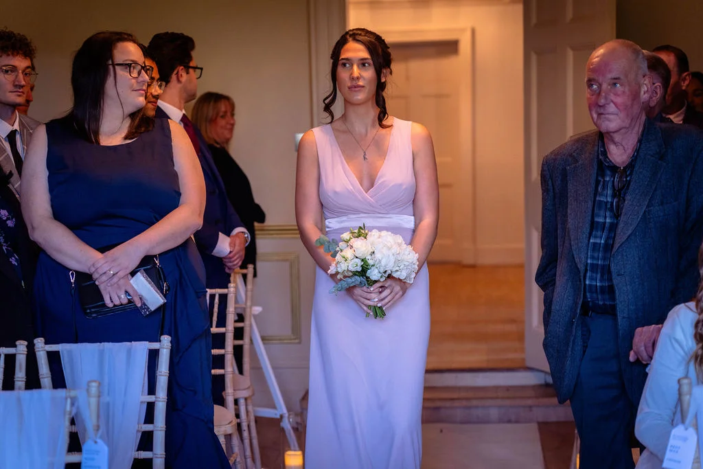 A bridesmaid in a pale pink dress holding a white bouquet stands near guests inside a wedding venue during the ceremony.