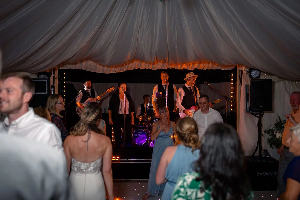 Wedding guests enjoy a lively evening reception inside a marquee with fabric-draped ceilings, watching a live band perform on stage.