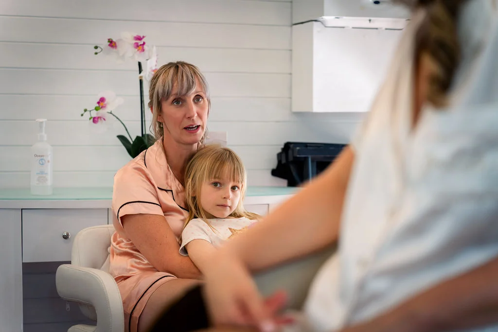 A woman and a young girl wearing matching pink pyjamas sit together indoors watching bridal preparations unfold.