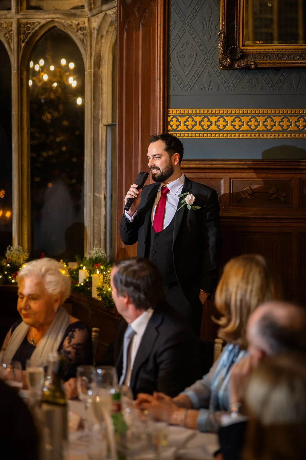 A groom delivers his wedding speech at the head table of an elegant country house reception, holding a microphone while guests listen attentively.