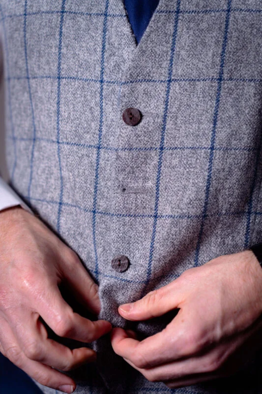 A close-up shot of a groom buttoning his grey tweed waistcoat featuring a blue check pattern during wedding morning preparations.