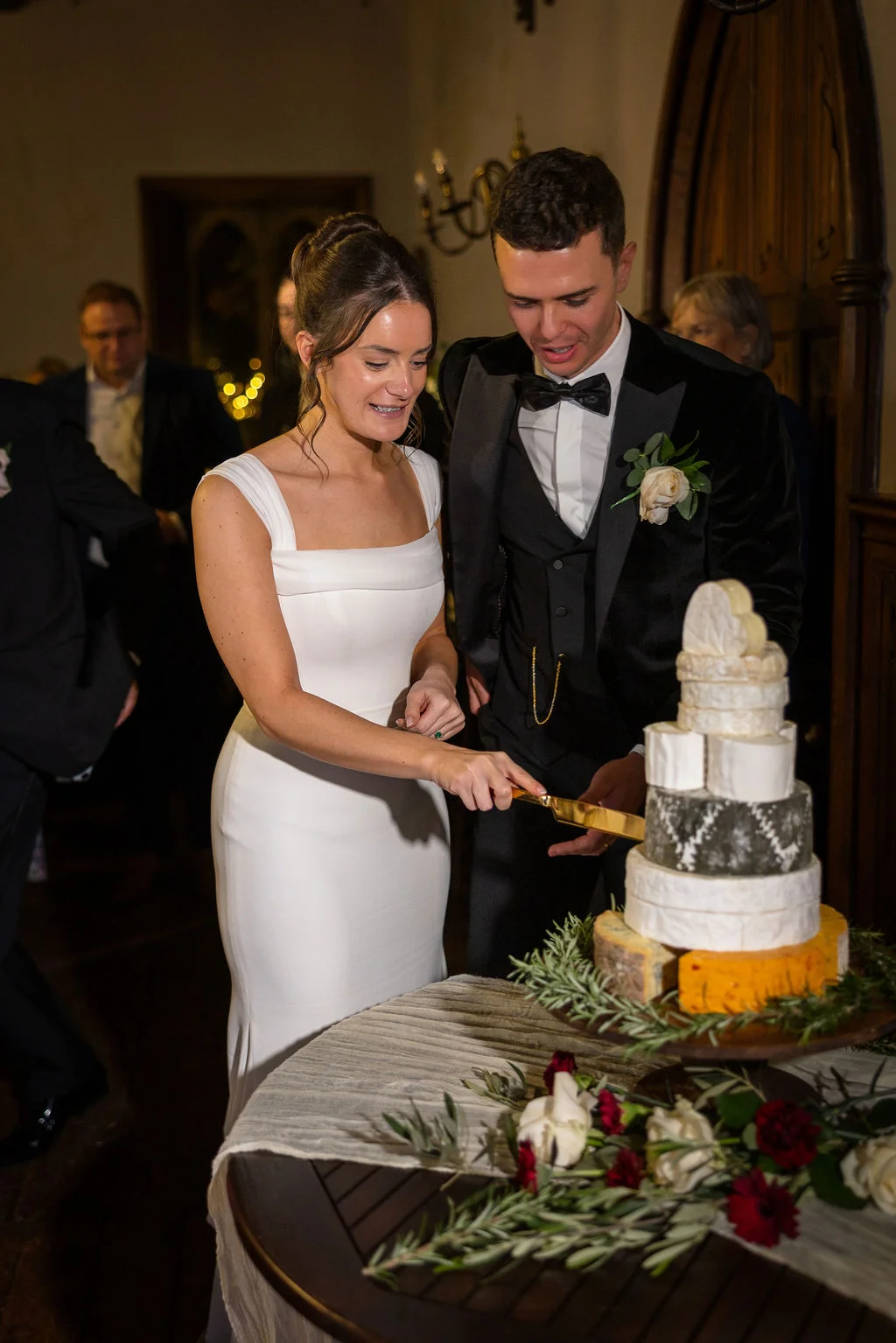 A smiling bride in a square-neckline white crepe gown and a groom in a black tuxedo cut a multi-tiered cheese wheel wedding cake adorned with rosemary.