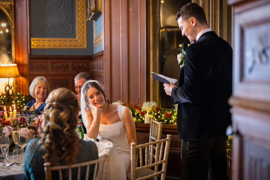 A smiling bride listens fondly as the groom delivers his speech during a warm, candlelit wedding reception in a wood-panelled dining room.