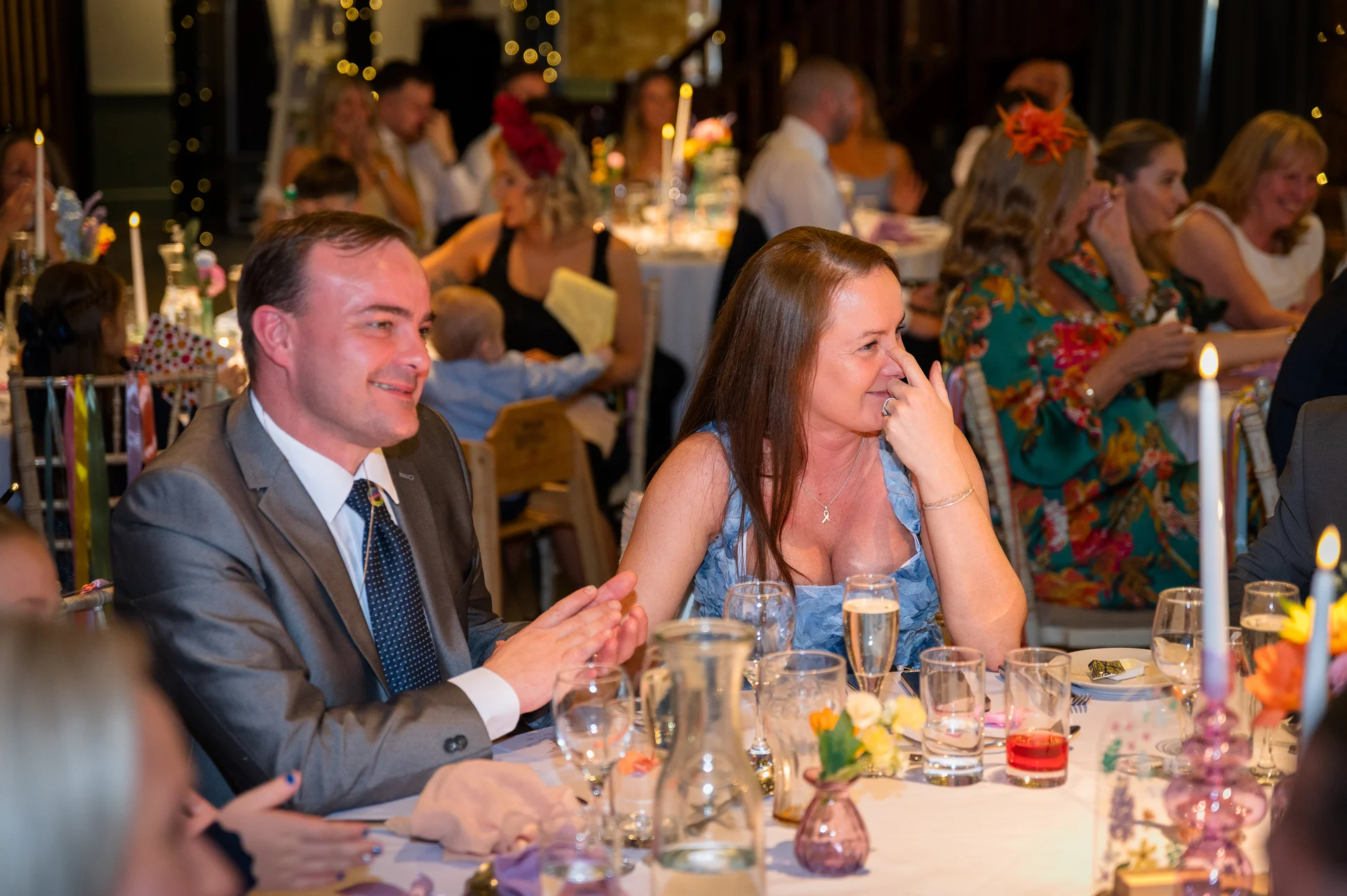 Wedding guests enjoy the speeches during the wedding breakfast, laughing and smiling around a beautifully set tablescape with taper candles and coloured glassware.