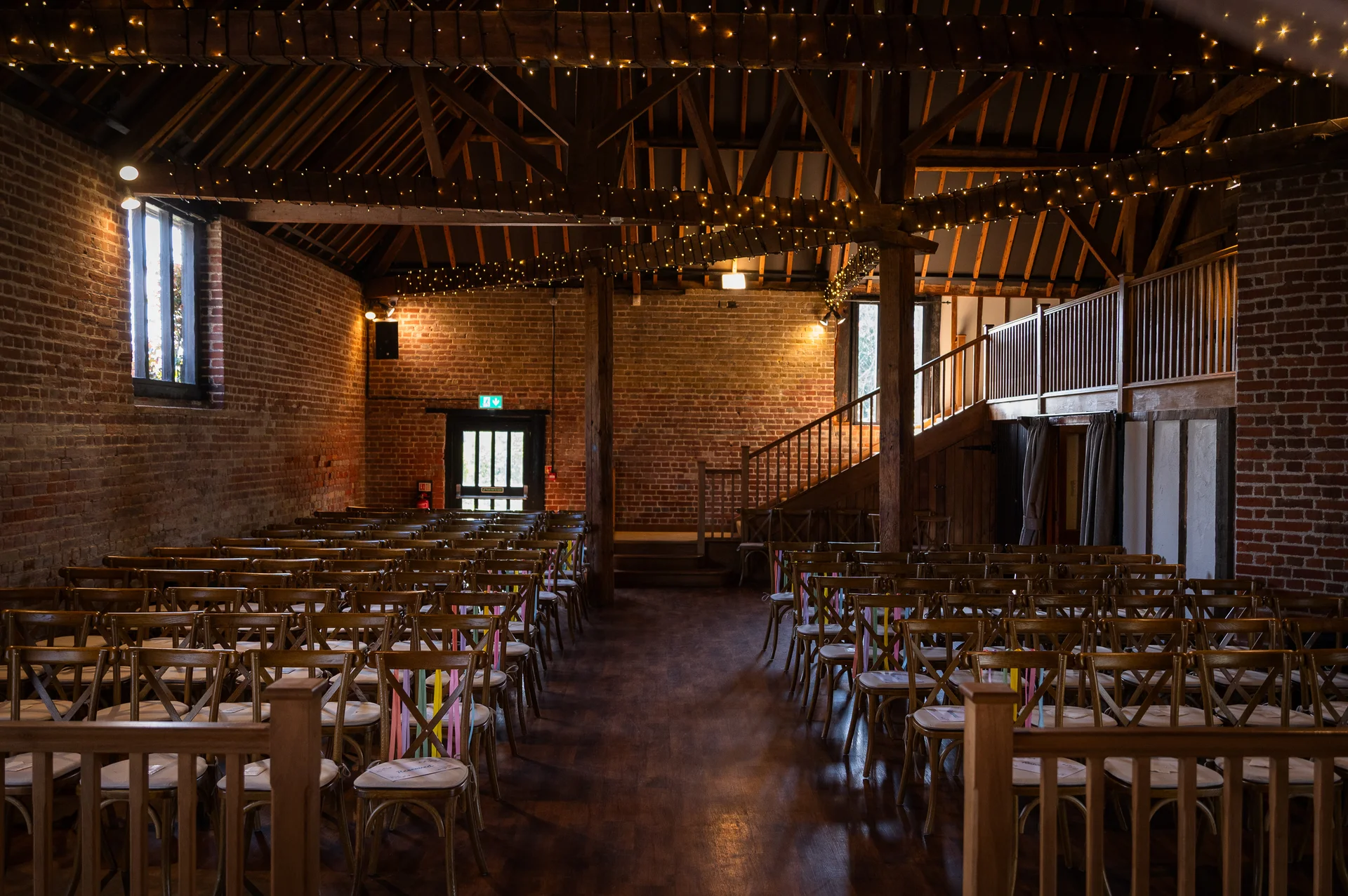 A rustic barn wedding venue set up for a ceremony with wooden chairs, exposed brick walls, and twinkling fairy lights draped across the wooden beams.