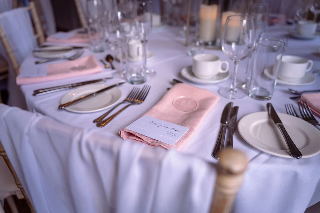 An elegantly set wedding reception table features crisp white linens, delicate blush pink napkins, polished cutlery, and glassware under soft natural light.