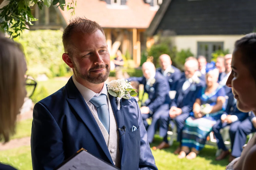 A groom looking lovingly toward his bride during an emotional outdoor wedding ceremony while guests watch from seating in the sunny background.