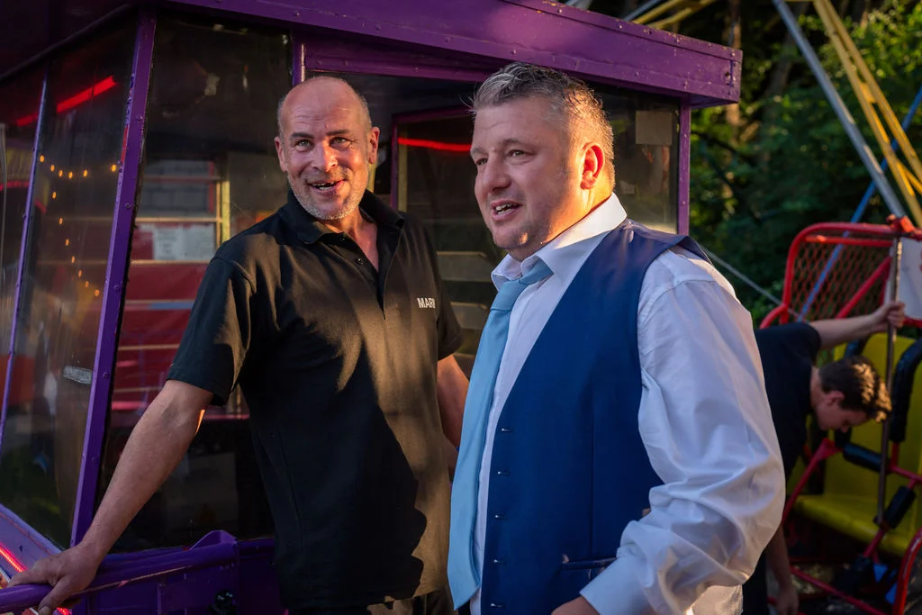 Two men smile warmly in the evening sun near a vibrant purple funfair attraction at a festival-style wedding.