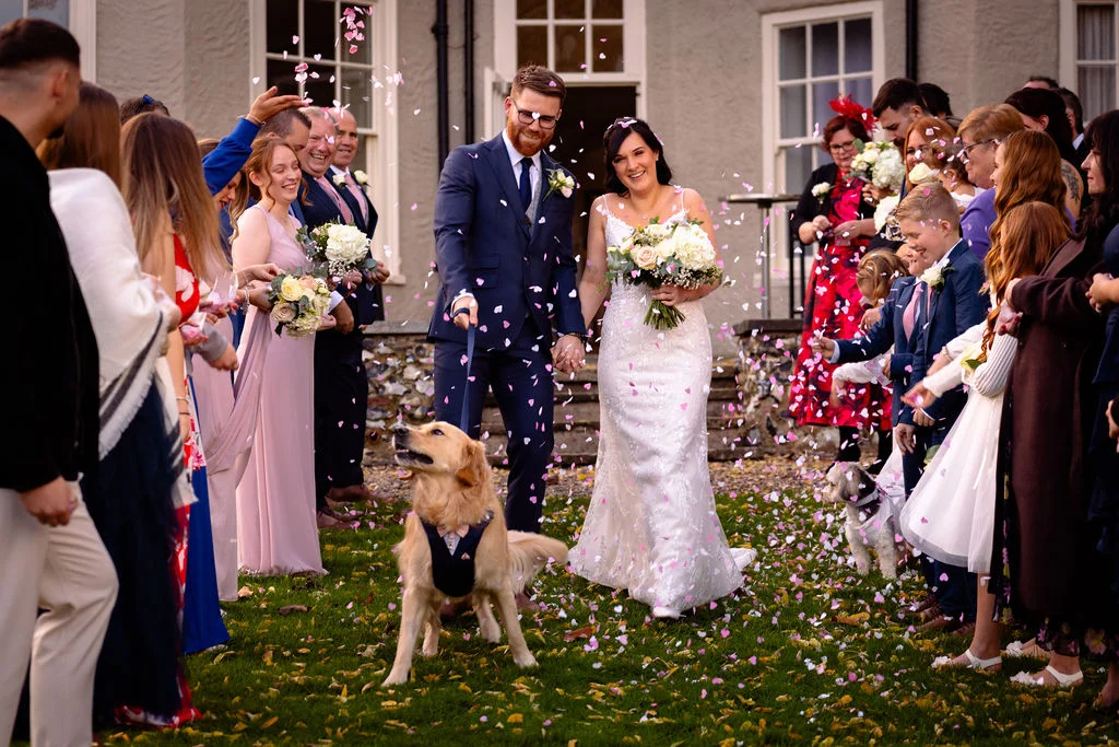 A smiling newlywed couple walks hand in hand through a cheering crowd of guests throwing confetti outside a country manor, accompanied by their golden retriever wearing a bow tie.