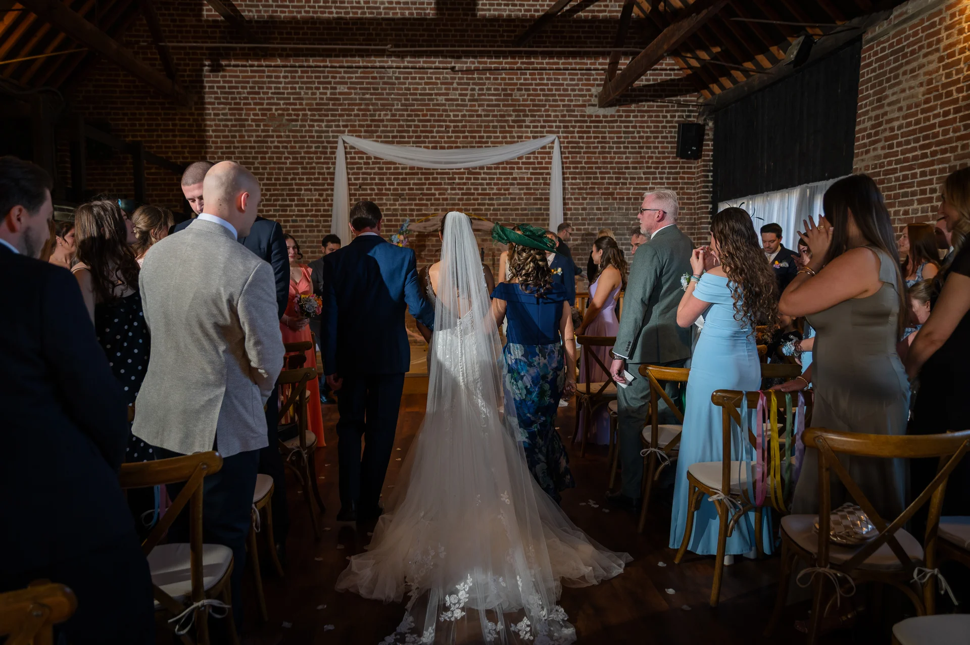 A bride in a lace gown with a cathedral veil walks down the aisle in a rustic brick barn venue with guests watching.