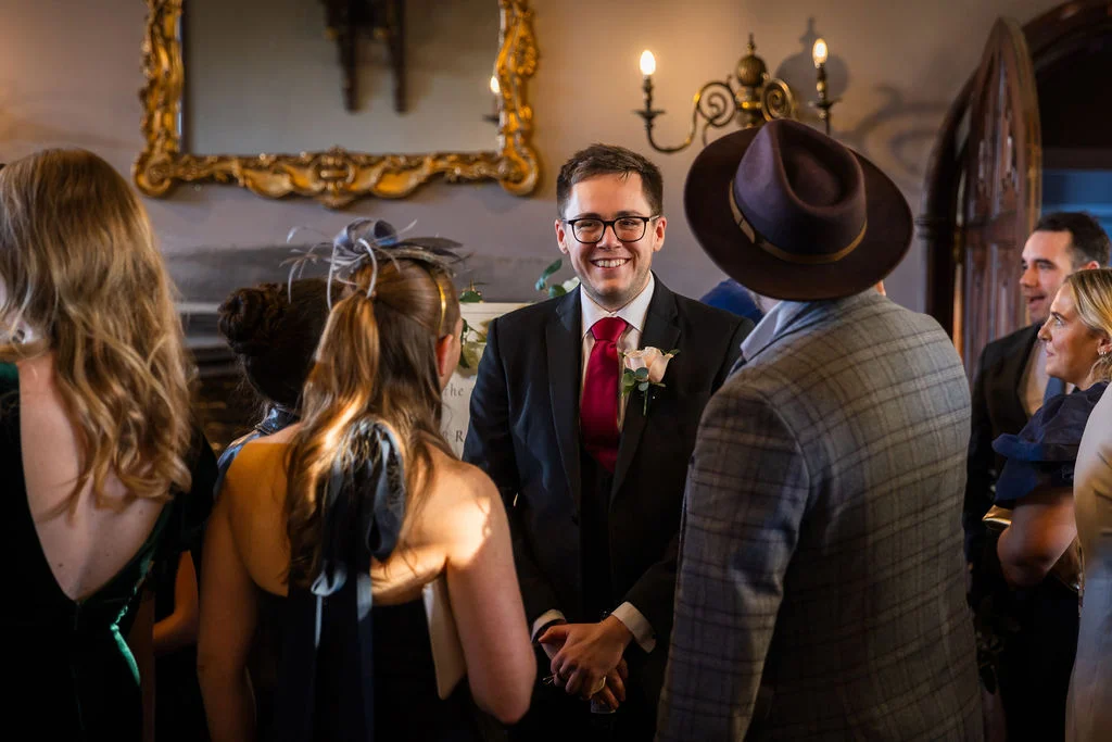A smiling groom wearing glasses and a dark suit with a red tie and boutonniere mingles with wedding guests in a classic country house reception room.