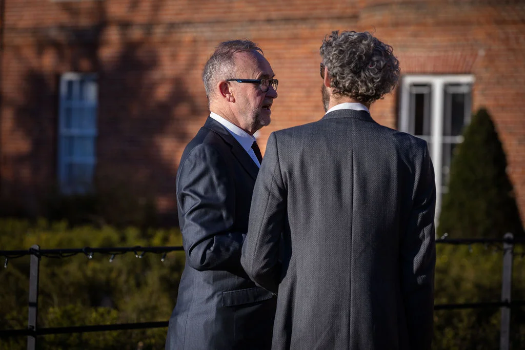 Two male wedding guests in grey suits converse outdoors against a red brick building during the drinks reception.