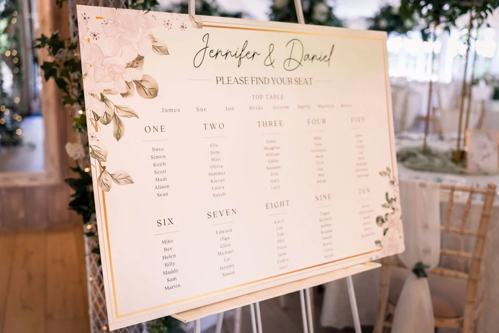 An elegant wedding seating chart displayed on a white easel featuring floral graphics and modern calligraphy to help guests find their tables.