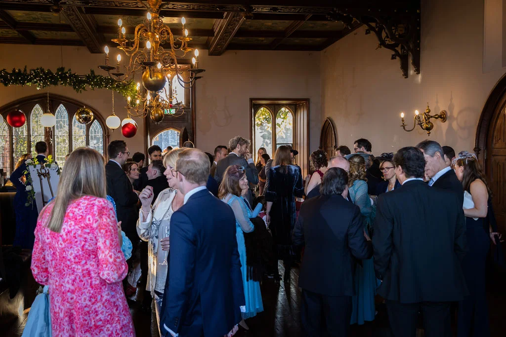 A bustling winter wedding drinks reception takes place inside a grand panelled manor house room illuminated by large chandeliers and decorated with festive baubles.