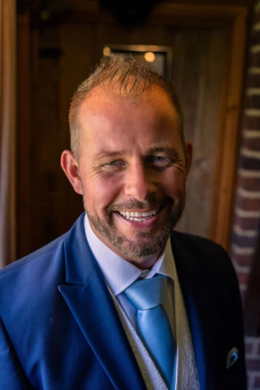 A smiling groom wearing a smart blue suit and matching blue tie poses indoors with a close-up portrait.