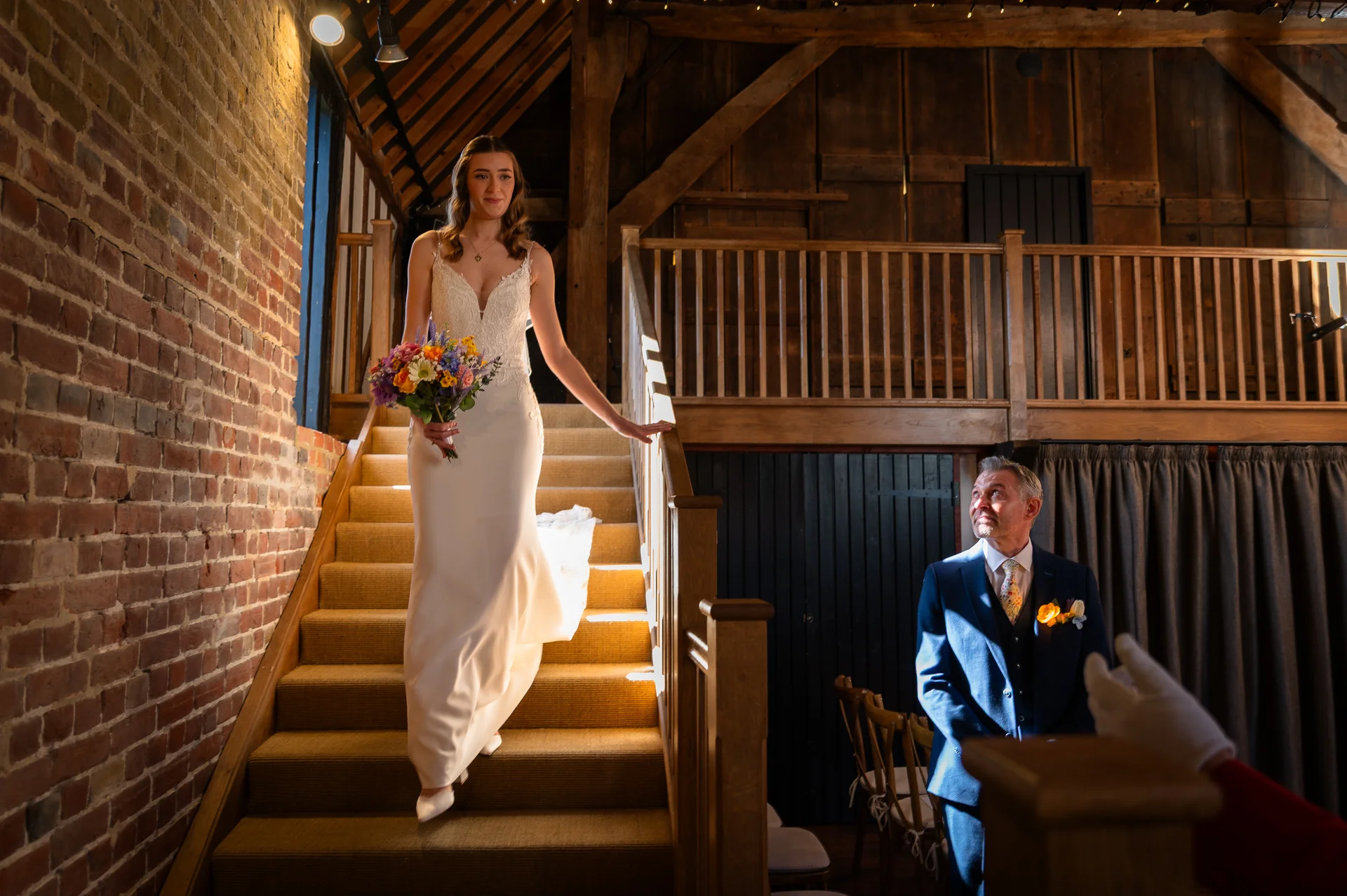 A smiling bride holding a colourful wildflower bouquet descends a wooden staircase inside an oak-framed rustic barn, while her father looks up at her with adoration.