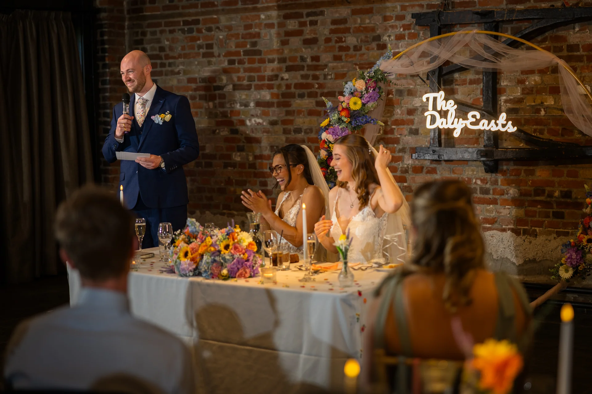 A groom delivers a speech at the top table in a rustic brick venue, making two smiling brides laugh as guests look on.