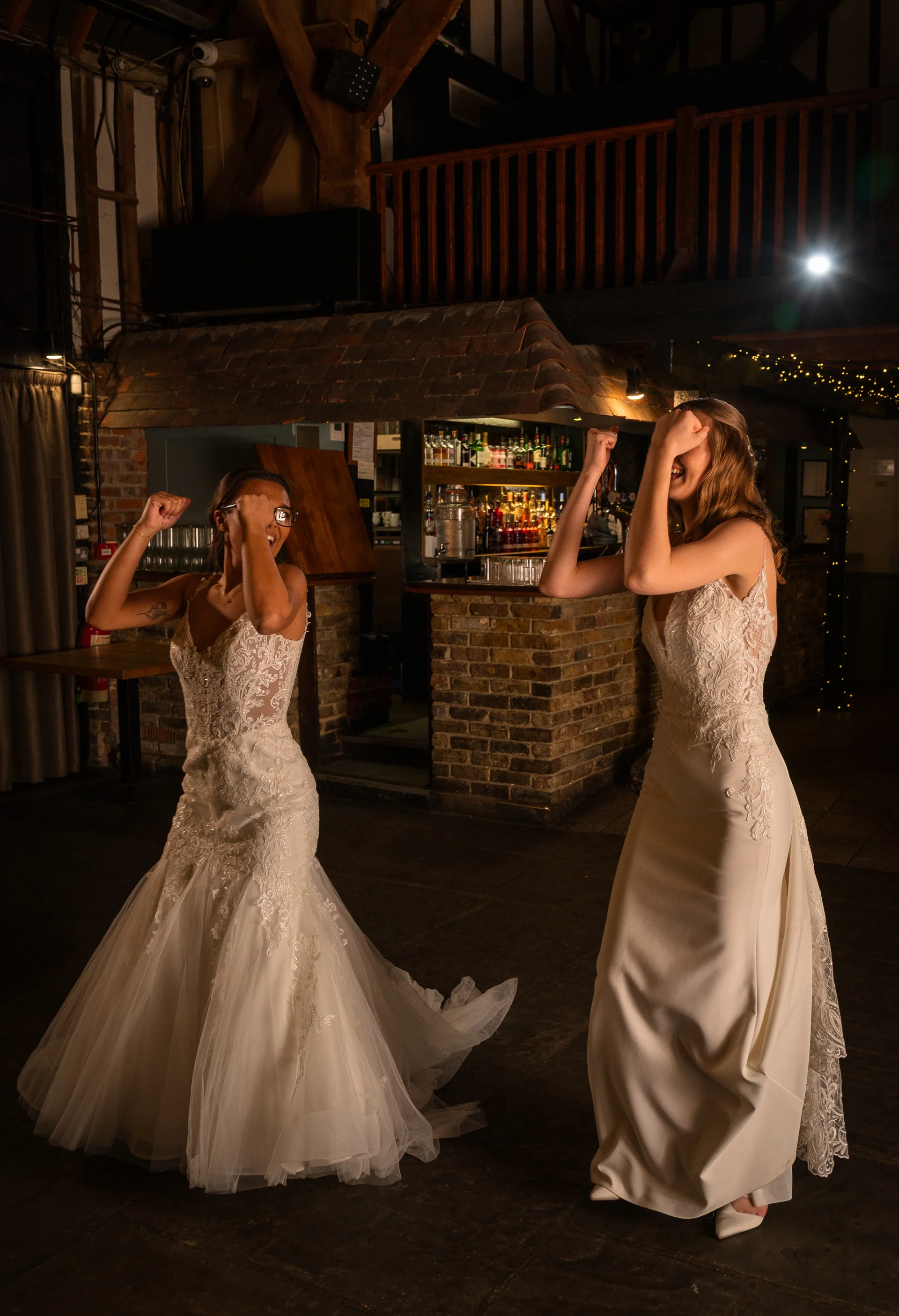 Two smiling brides in lace wedding dresses celebrate energetically on the dance floor inside an atmospheric barn venue with exposed brickwork.