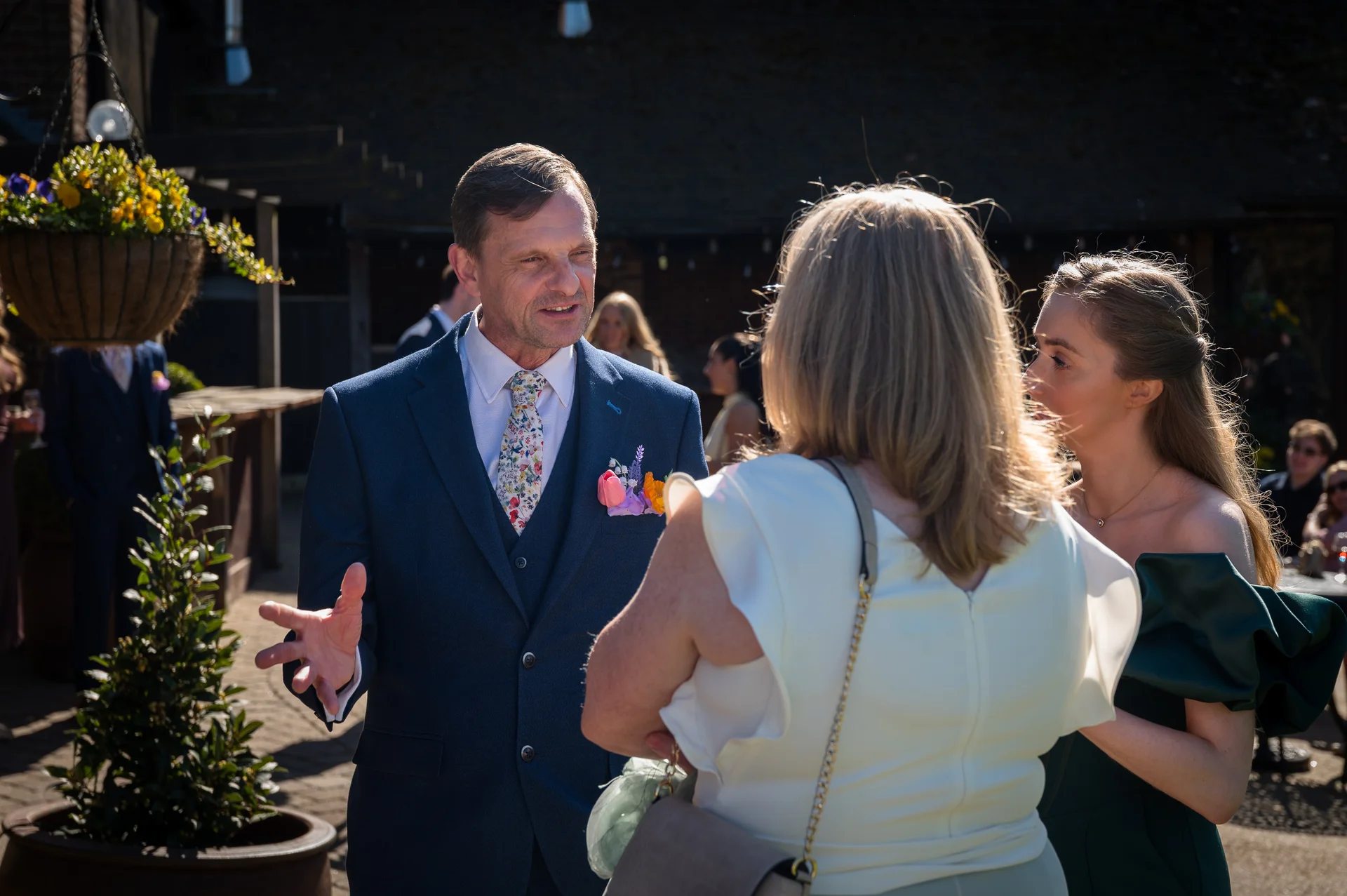 A well-dressed man in a navy suit gestures enthusiastically while chatting with guests outdoors on a sunny wedding day.