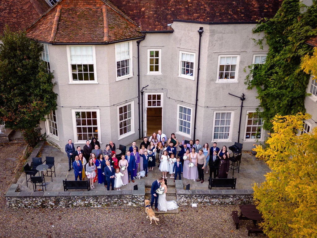An aerial drone shot captures a large wedding party gathered in the courtyard of a country house surrounded by autumn foliage.