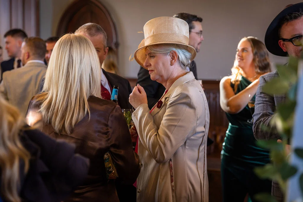 Well-dressed wedding guests mingle and converse during the drinks reception, with a woman in a stylish cream outfit and hat in profile.
