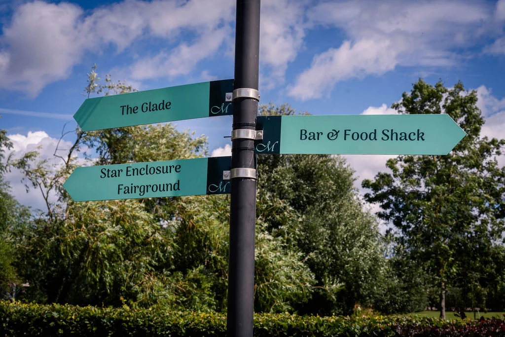 A teal wooden wedding signpost with three directional arrows pointing towards The Glade, Bar and Food Shack, and Star Enclosure Fairground under a bright blue sky with fluffy white clouds.