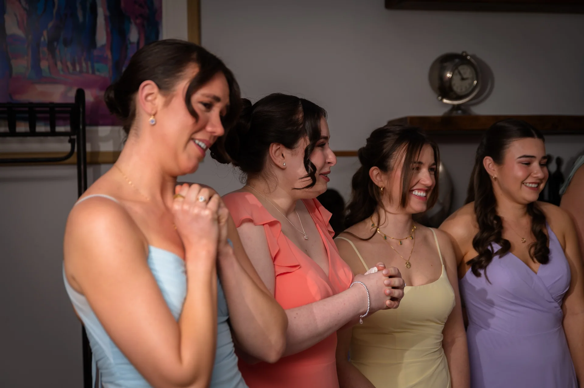 A group of four smiling and laughing bridesmaids wearing pastel-coloured dresses in shades of blue, peach, yellow, and purple.