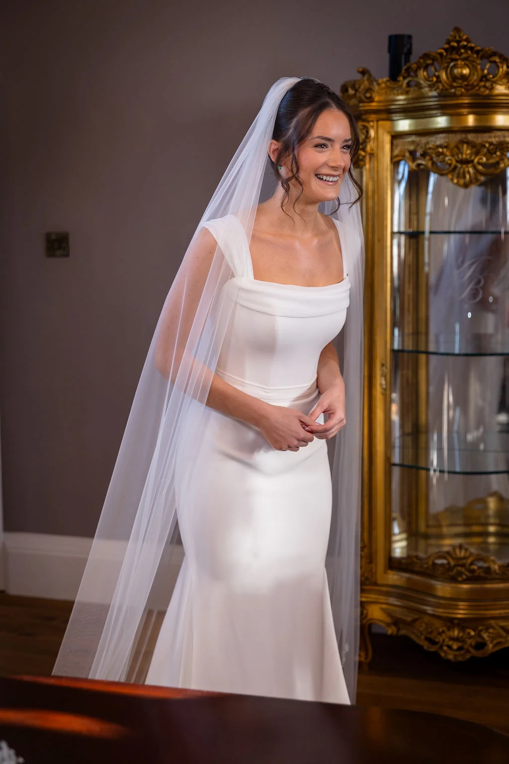 A smiling bride wearing a modern square-neck crepe gown and a long veil stands indoors next to a gold ornate cabinet.