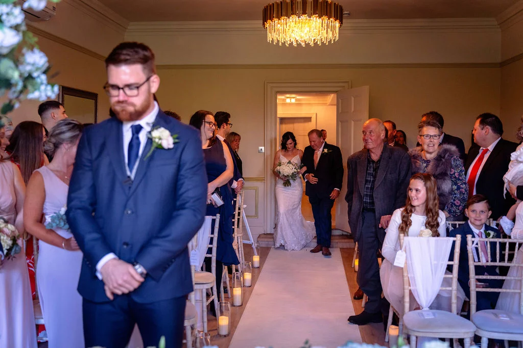 A groom in a navy suit stands facing forward at the altar while the bride walks down the aisle accompanied by her father.