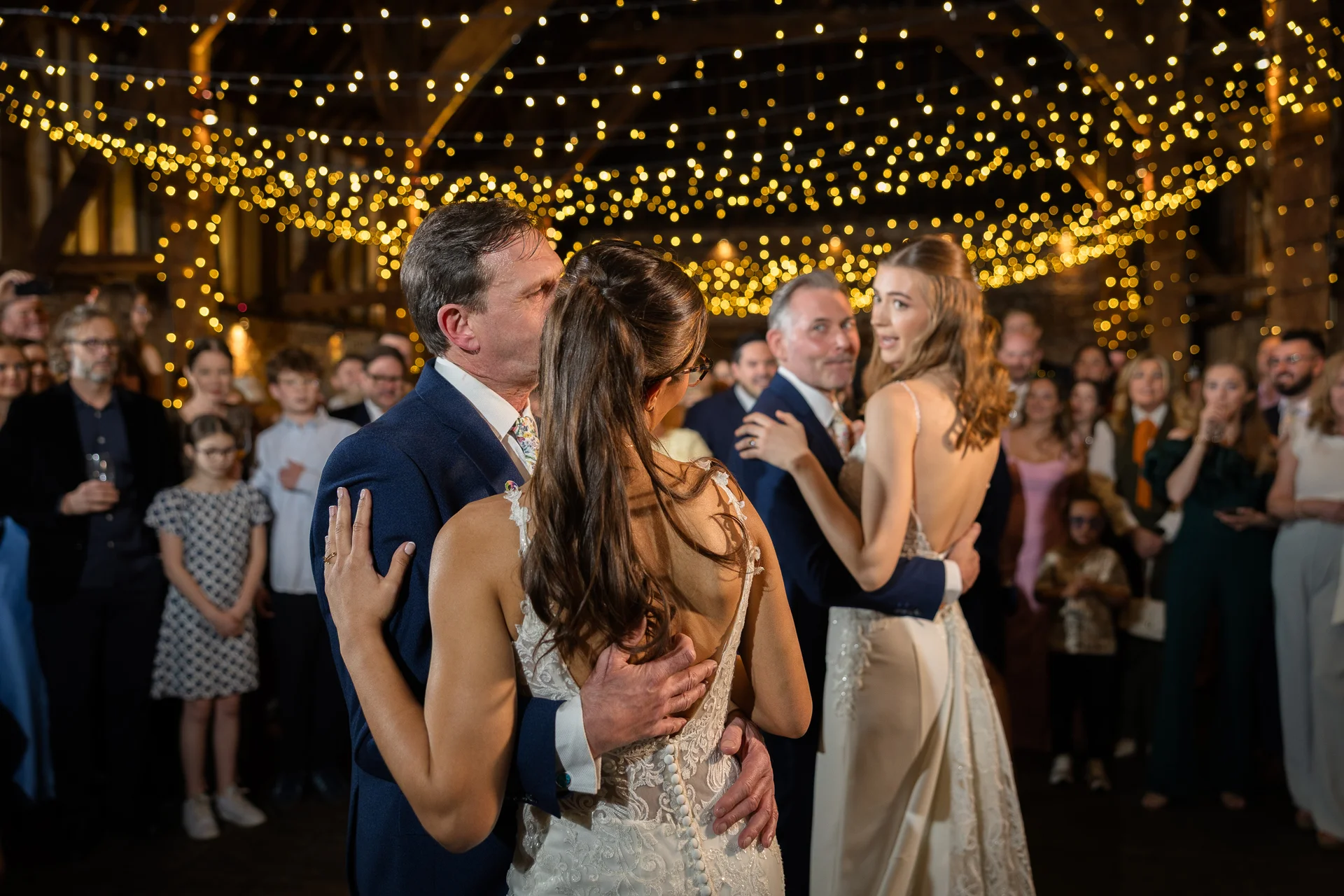 A father and bride share a tender dance during the evening reception beneath a magnificent canopy of twinkling fairy lights inside a rustic oak-framed barn.