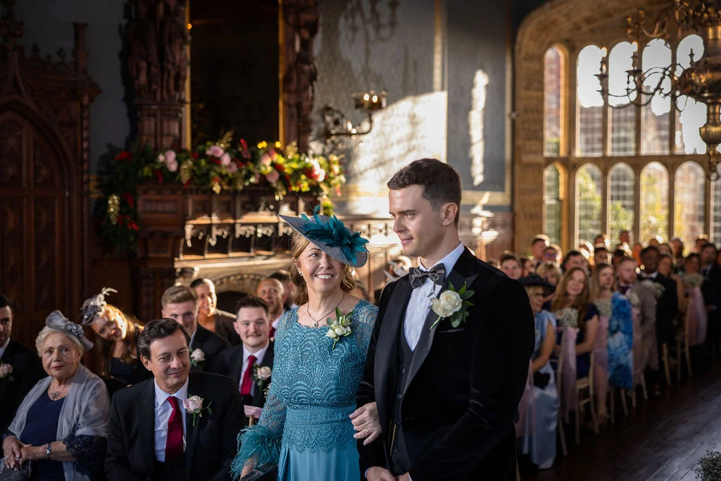 A groom in a black velvet tuxedo and a guest in a teal lace dress walk down the aisle in a grand country house with large windows and a decorated fireplace.