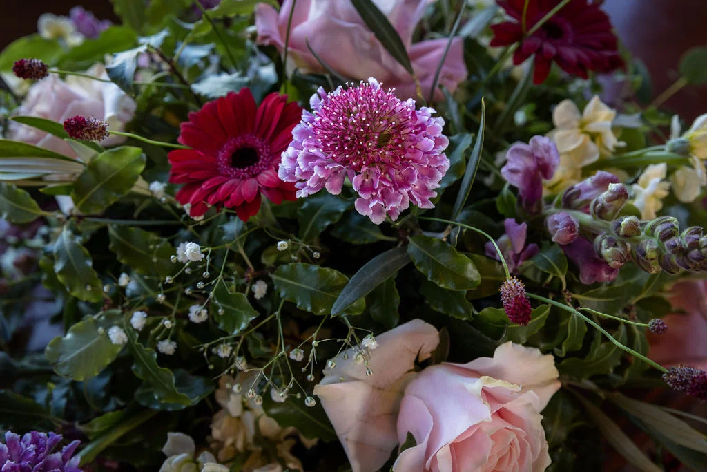 A close-up view of a lush, hand-tied wedding bouquet featuring pale pink roses, deep red gerbera daisies, and delicate scabiosa flowers amidst greenery.