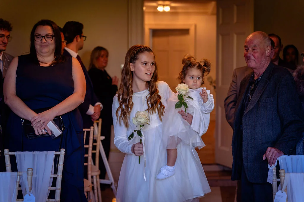 A young bridesmaid holding a white rose and carrying a toddler flower girl during the wedding ceremony.