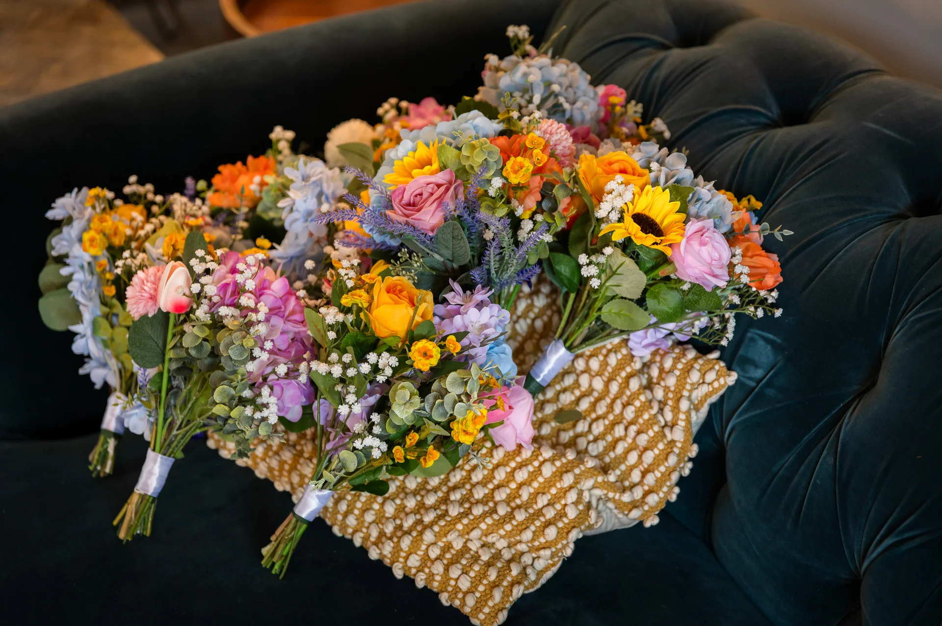 A colourful collection of hand tied wedding bouquets featuring sunflowers, pink roses, and baby's breath rests on textured gold cushions atop a dark velvet sofa.