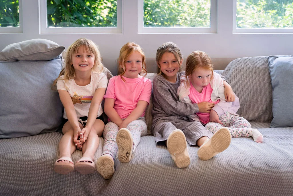 Four young girls sit together smiling on a large grey sofa bathed in soft window light.